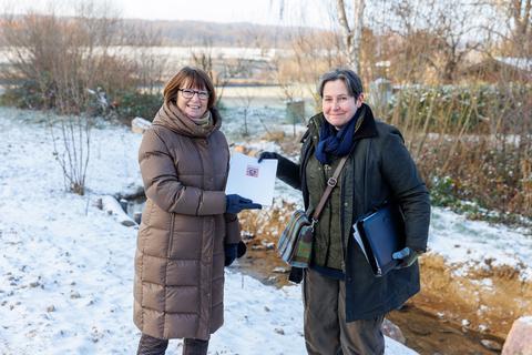 Die hessische Umweltministerin Priska Hinz (links) übergibt am Auringer Wickerbach Bescheide über Fördergelder an Stadträtin Bettina Gies.